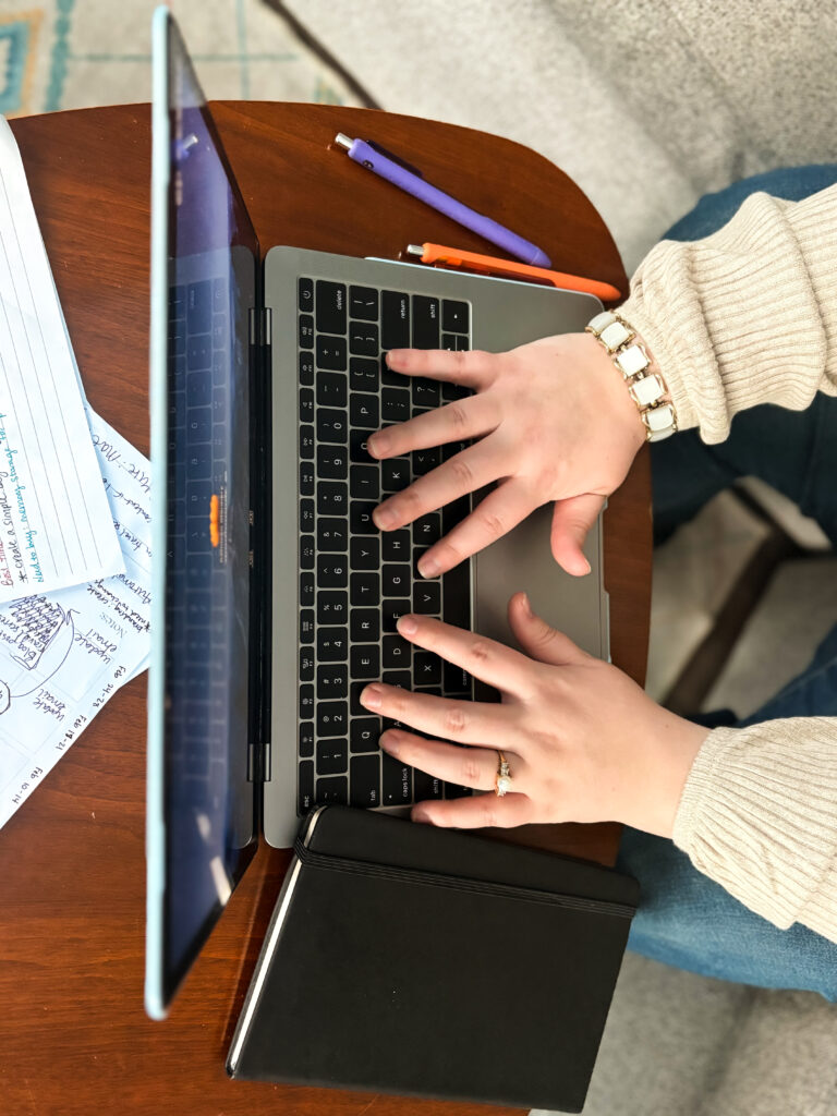 Kathleen typing on a laptop keyboard. The laptop is placed on a wooden coffee table, alongside a black notebook, handwritten notes, and two pens. The setting suggests a focused and productive work-from-home or study session.